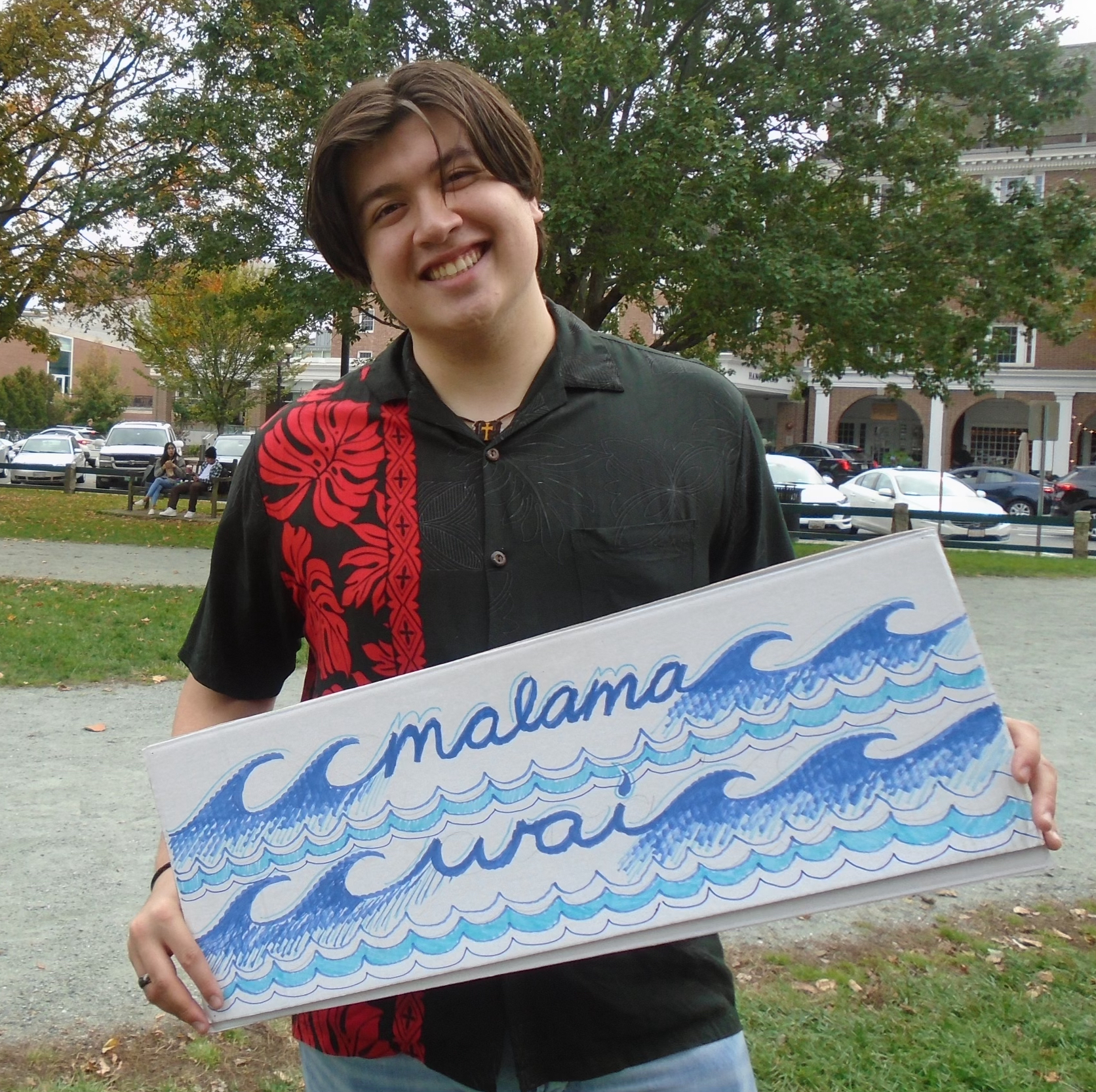 Gabriel H. Gilbert wearing red and black aloha shirt holding a sign that reads "malama wai" on the Dartmouth Green, 2022.